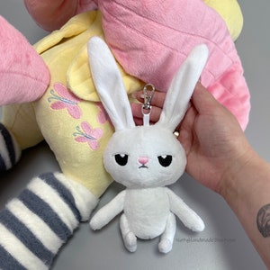 May include: White plush bunny keychain with black eyes and a pink nose. The bunny is sitting with its arms and legs outstretched. The keychain has a silver metal ring and clip. The bunny is attached to a larger plush toy with pink and yellow fabric.