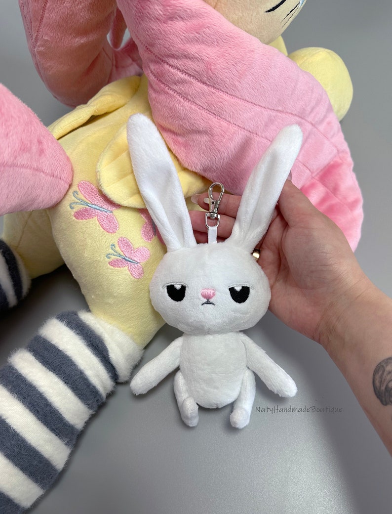 May include: White plush bunny keychain with black eyes and a pink nose. The bunny is sitting with its arms and legs outstretched. The keychain has a silver metal ring and clip. The bunny is attached to a larger plush toy with pink and yellow fabric.