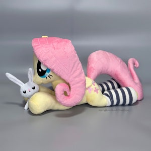 May include: A plush pink and yellow pony with a pink mane and tail, wearing black and white striped socks, is lying down with a white plush bunny in its arms. The pony has a blue eye and a pink nose. The bunny has a pink nose and black eyes.