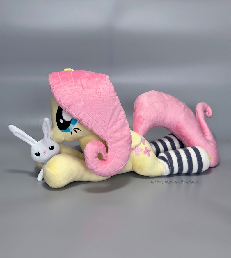 May include: A plush pink and yellow pony with a pink mane and tail, wearing black and white striped socks, is lying down with a white plush bunny in its arms. The pony has a blue eye and a pink nose. The bunny has a pink nose and black eyes.