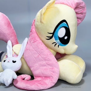 May include: A plush toy of Pinkie Pie from My Little Pony, in a light yellow and pink color scheme, with a white bunny plush toy in front of her.