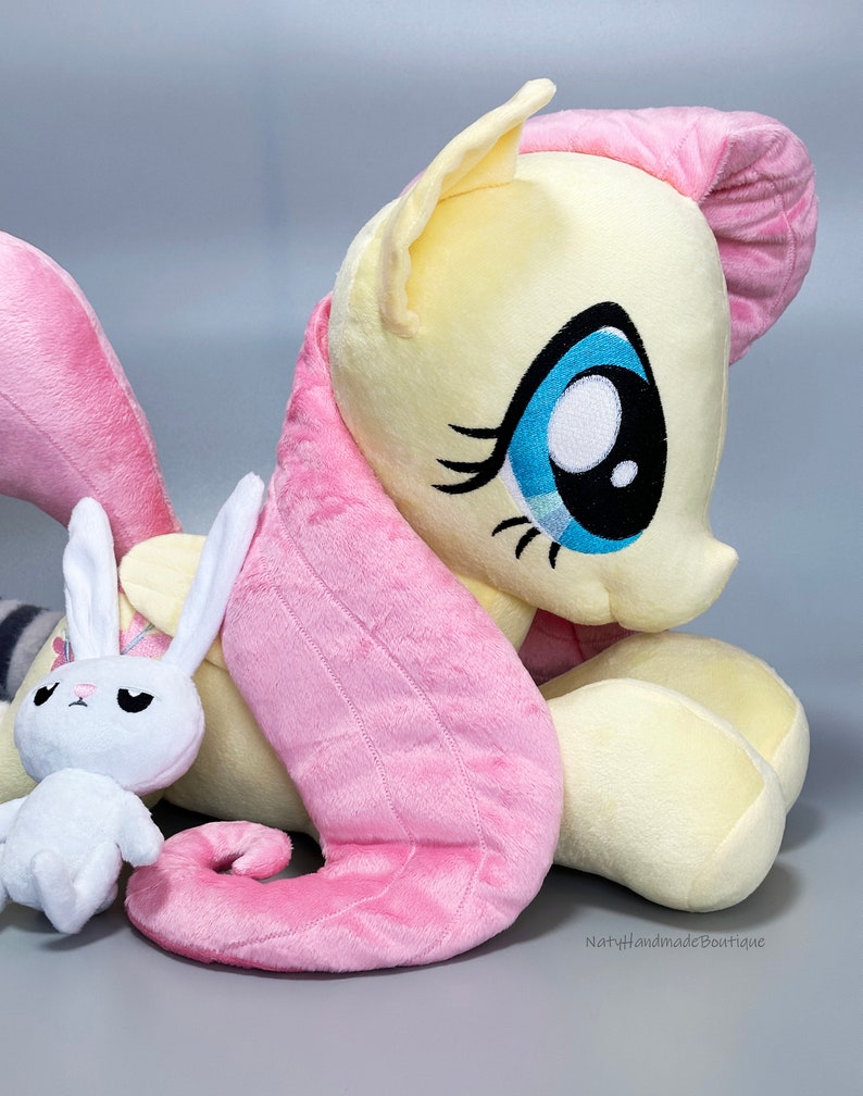 May include: A plush toy of Pinkie Pie from My Little Pony, in a light yellow and pink color scheme, with a white bunny plush toy in front of her.