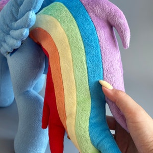 May include: A plush toy of a blue pony with a rainbow-colored tail. The tail is made of soft, colorful fabric and is shaped like a rainbow. The pony is standing on a light gray background.