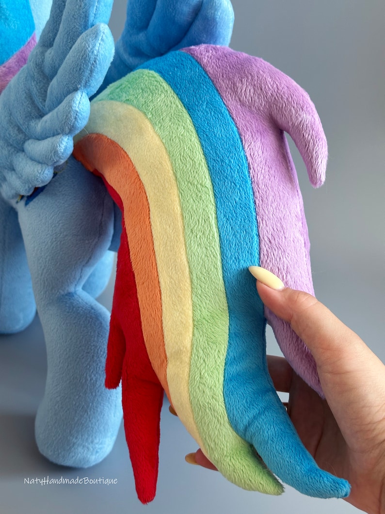 May include: A plush toy of a blue pony with a rainbow-colored tail. The tail is made of soft, colorful fabric and is shaped like a rainbow. The pony is standing on a light gray background.