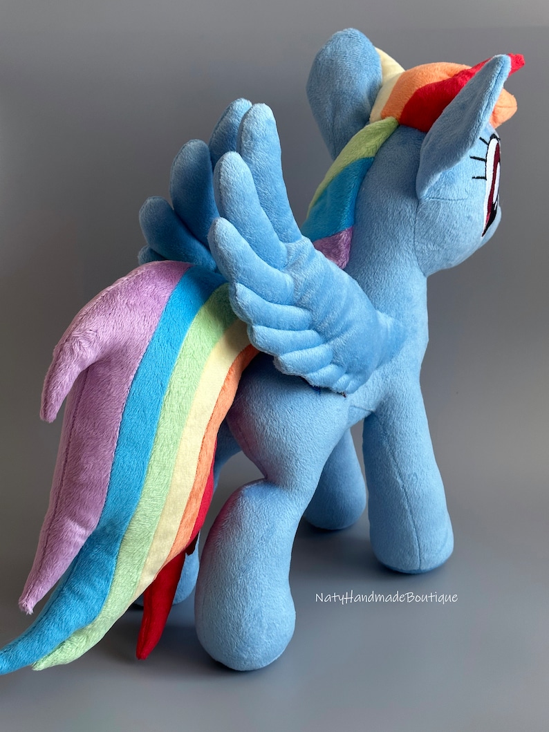 May include: A plush toy of Rainbow Dash from My Little Pony. The toy is blue with a rainbow mane and tail. It has wings and is standing on a gray surface. The text "NatyHandmadeBoutique" is visible in the bottom right corner.