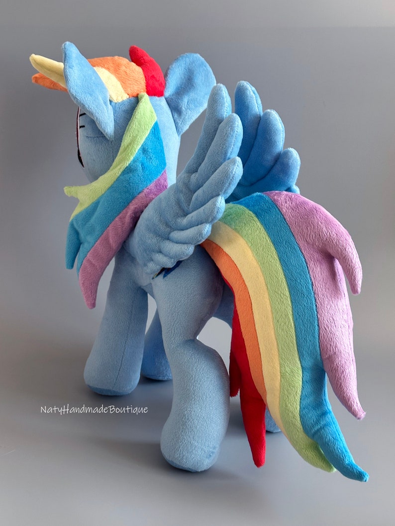 May include: A plush toy of Rainbow Dash from My Little Pony. The toy is blue with a rainbow mane and tail. The toy is standing on a gray surface.