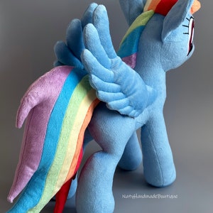 May include: A plush toy of Rainbow Dash from My Little Pony. The toy is blue with a rainbow mane and tail. It has wings and is standing on a gray surface. The text "NatyHandmadeBoutique" is visible in the bottom right corner.