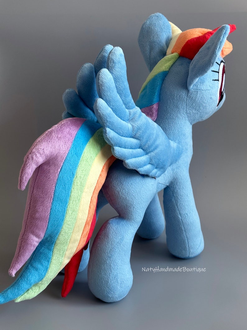 May include: A plush toy of Rainbow Dash from My Little Pony. The toy is blue with a rainbow mane and tail. It has wings and is standing on a gray surface. The text "NatyHandmadeBoutique" is visible in the bottom right corner.