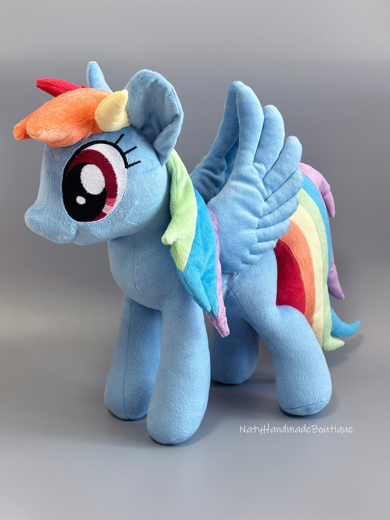 May include: A plush toy of Rainbow Dash from My Little Pony. The toy is light blue with a rainbow mane and tail. It has a pink nose, red eyes, and a yellow horn. The toy is standing on a gray surface.