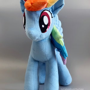 May include: A blue plush toy pony with a rainbow mane and tail. The pony has large, red eyes and a white horn. The toy is standing on a gray background.