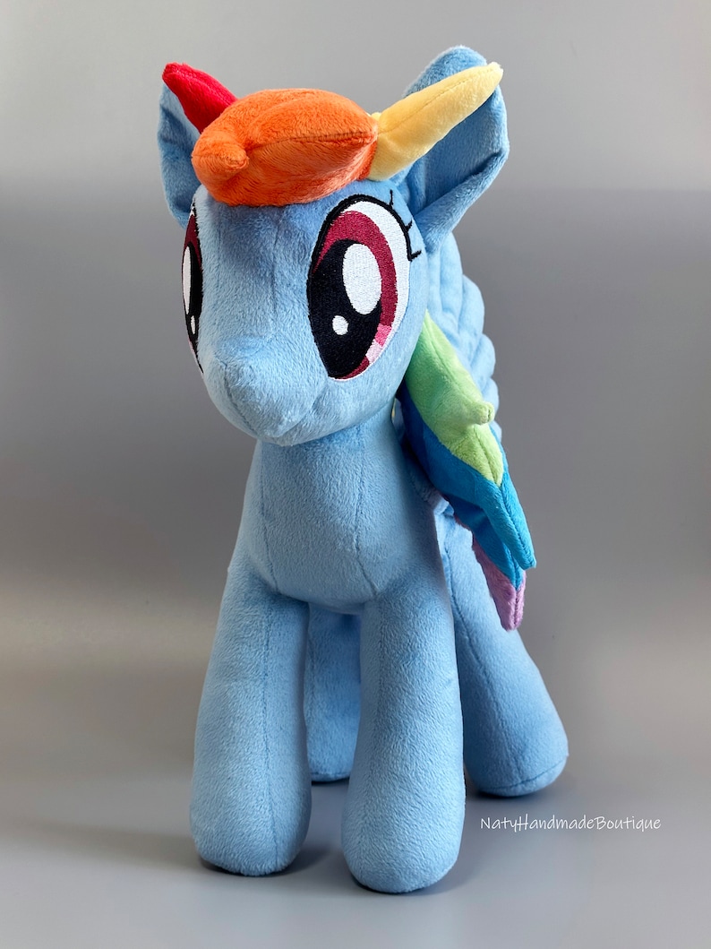 May include: A blue plush toy pony with a rainbow mane and tail. The pony has large, red eyes and a white horn. The toy is standing on a gray background.