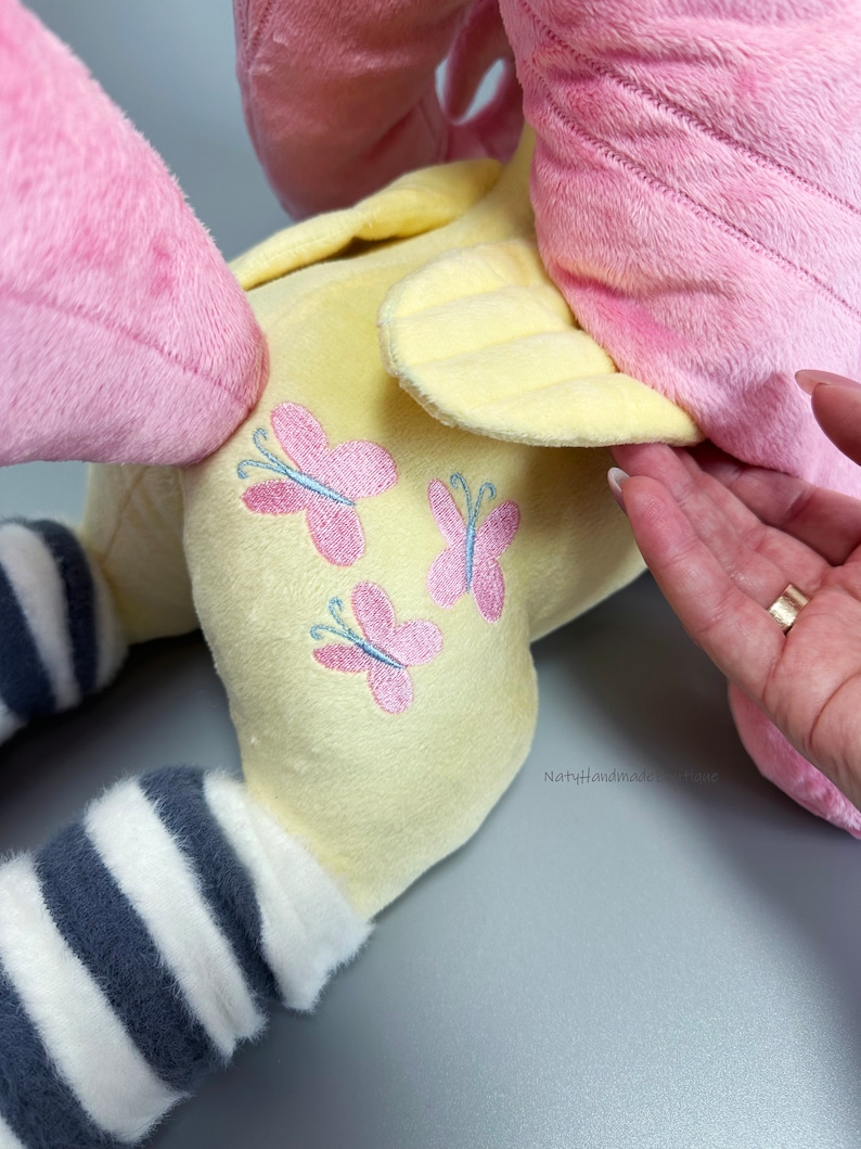 May include: A plush toy with pink and yellow fur, a striped leg, and embroidered pink butterflies on the back. The toy is made by NattyHandmade.