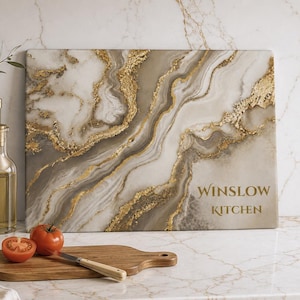 May include: A rectangular cutting board featuring a marbled pattern in shades of white, beige, and gray with gold-colored accents. The text "WINSLOW KITCHEN" is printed in gold on the lower right side of the board, which has a smooth surface.