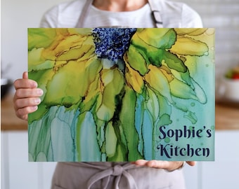 Peut inclure: Un plan de travail rectangulaire avec un motif de tournesol à l'aquarelle dans des tons de jaune, vert et bleu. Le texte "Sophie's Kitchen" est affiché en écriture bleu foncé. La planche est tenue par une personne.