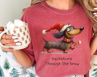 May include: Crimson t-shirt with a cartoon dachshund wearing a Santa hat and scarf, and the text "Dachshund Through The Snow." The dog is decorated with Christmas lights. The shirt is worn with blue jeans.