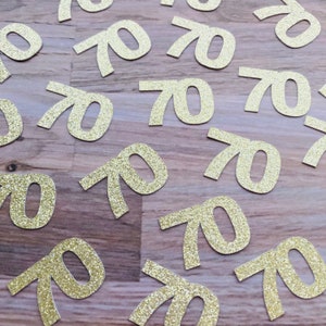 May include: Gold glitter confetti with the number 70 printed on each piece. The confetti is scattered on a wooden surface.