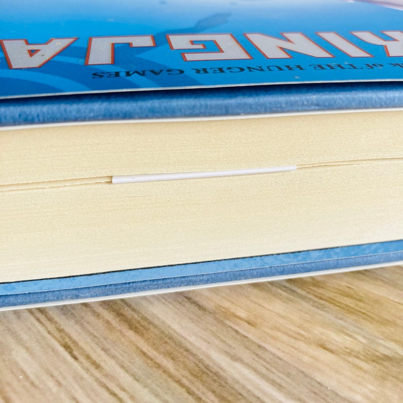 以下が含まれることがあります： 青い表紙の書籍の背表紙のクローズアップ。白い文字で「The Hunger Games」と書いてあります。本は閉じられており、背表紙は少し傾いています。