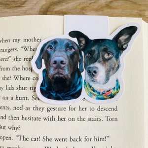 May include: Two dog-shaped bookmarks. One features a brown Labrador Retriever with a blue scarf, the other a black and white dog with a colorful scarf. The bookmarks are placed on the pages of a book.