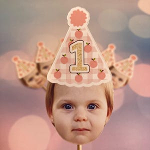Peut inclure: Un chapeau d'anniversaire en vichy rose et blanc avec un motif de p&ecirc;che et le chiffre "1" en paillettes dor&eacute;es. Le chapeau est sur un b&acirc;ton et a un visage d'enfant en arri&egrave;re-plan.
