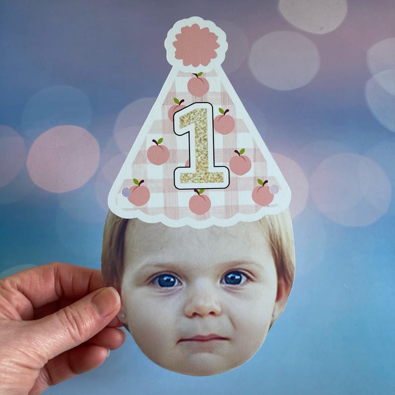 Peut inclure: Un chapeau de f&ecirc;te en vichy rose et blanc avec un motif de p&ecirc;che et un num&eacute;ro 1 en paillettes dor&eacute;es. Le chapeau est port&eacute; par un enfant aux cheveux blonds et aux yeux bleus.