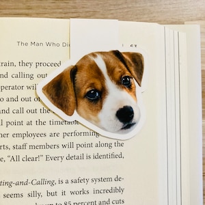May include: A bookmark featuring a Jack Russell Terrier puppy's face, with brown and white markings, is placed on the pages of a book. The bookmark is a cut-out of the dog's head, with a white border.