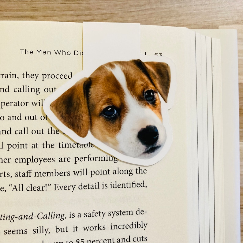 May include: A bookmark featuring a Jack Russell Terrier puppy's face, with brown and white markings, is placed on the pages of a book. The bookmark is a cut-out of the dog's head, with a white border.