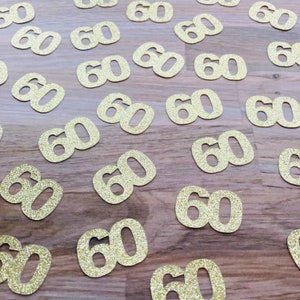 May include: Gold glitter confetti with the number 60 printed on each piece. The confetti is scattered on a wooden surface.