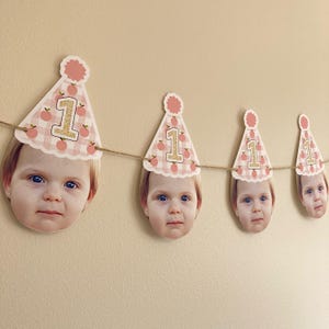 Peut inclure: Une banni&egrave;re d'anniversaire rose et blanche avec des motifs de p&ecirc;che et le chiffre "1" sur chaque drapeau. Chaque drapeau a une photo du visage d'un b&eacute;b&eacute; dans un chapeau de f&ecirc;te.