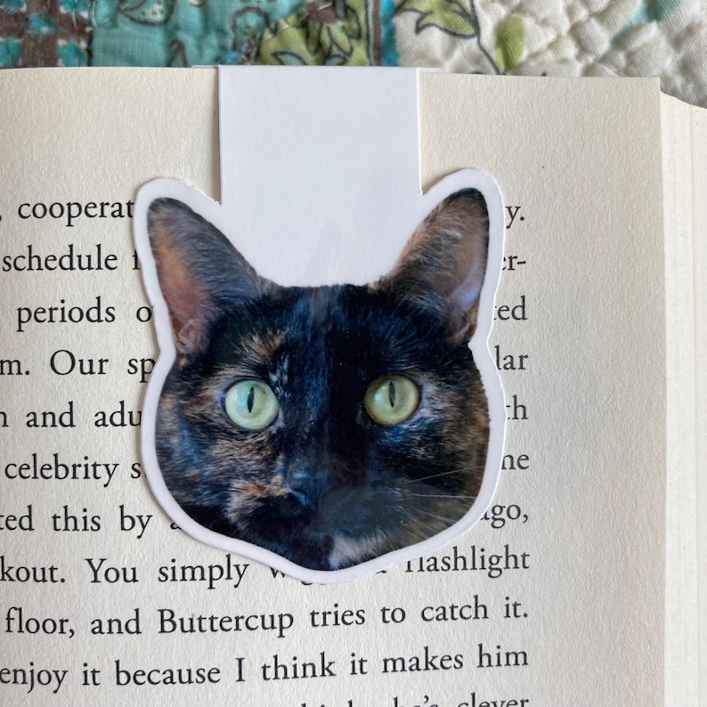May include: A cat-shaped bookmark featuring a close-up of a tortoiseshell cat's face with green eyes. The bookmark is placed in a book with printed text. The cat's fur is a mix of black, brown, and tan.