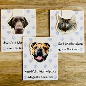 May include: Three magnetic bookmarks featuring animal faces: a dog, a cat, and a bulldog. Each bookmark has a white tab and is attached to a card with paw prints and hearts. The text "Heartfelt Marketplace Magnetic Bookmark" is printed on each card.