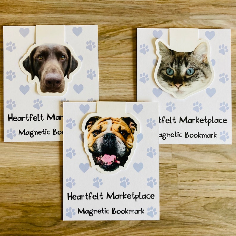 May include: Three magnetic bookmarks featuring animal faces: a dog, a cat, and a bulldog. Each bookmark has a white tab and is attached to a card with paw prints and hearts. The text "Heartfelt Marketplace Magnetic Bookmark" is printed on each card.