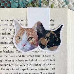 May include: A white bookmark featuring two cat faces. One cat is orange and white, the other is calico with black, brown, and orange fur. The bookmark is placed in an open book with printed text.