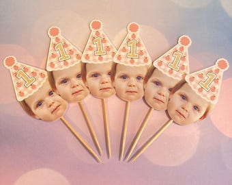 Op de afbeelding: Zes cupcake toppers met een babygezicht en een feesthoedje met het nummer "1". De hoeden hebben een perzik- en wit ruitpatroon en een perzik-kleurige pompon.