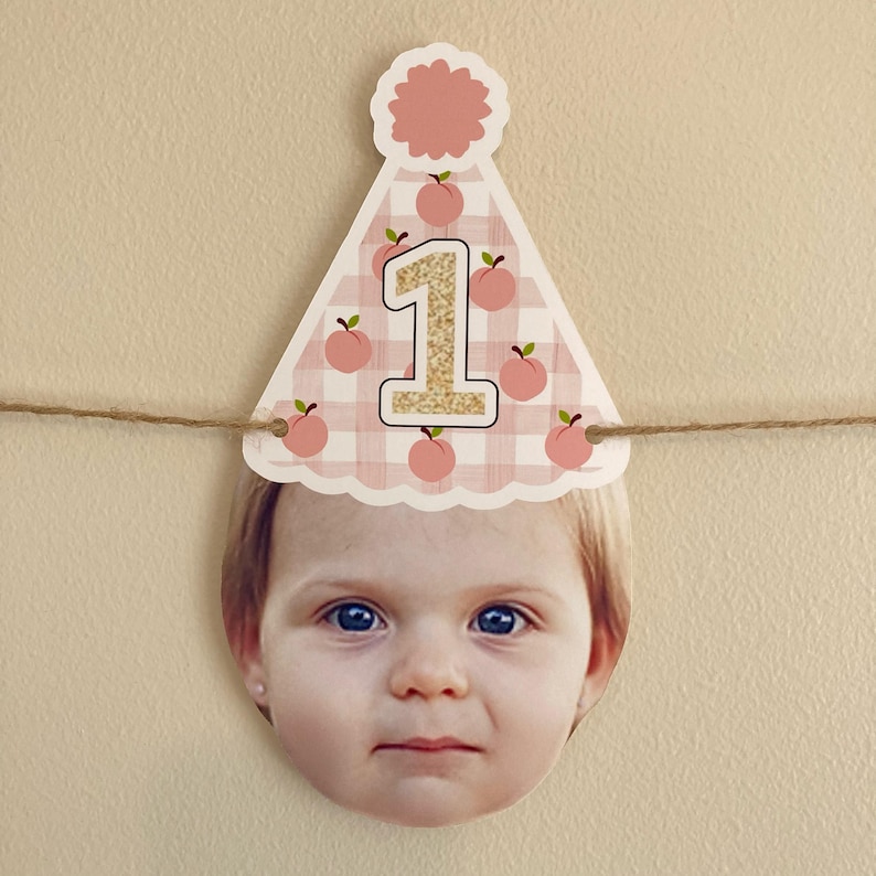 Peut inclure: Un chapeau de f&ecirc;te en vichy rose et blanc avec des illustrations de p&ecirc;ches et un num&eacute;ro "1" en paillettes dor&eacute;es sur le devant. Le chapeau est attach&eacute; &agrave; une ficelle et a un visage d'enfant d&eacute;coup&eacute; dans le bas.