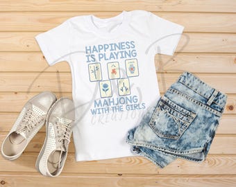 Könnte beinhalten: Weißes T-Shirt mit dem Text "Happiness is playing Mahjong with the girls" und Mahjong-Kachel-Grafiken. Das Shirt wird mit Jeansshorts und silbernen Turnschuhen auf einer Holzoberfläche kombiniert.