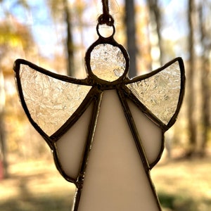 May include: A white stained glass angel ornament with a frosted glass effect. The angel has a halo and wings. The ornament is hanging from a string.