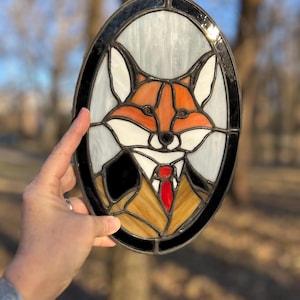 May include: A stained glass window depicting a fox wearing a brown suit and a red tie. The fox has orange fur and white facial markings. The window is oval shaped and has a black frame.