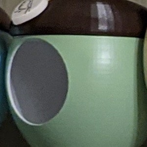 May include: A whimsical, acorn-shaped birdhouse. The birdhouse is painted in a light green color with a dark brown top. A circular opening is visible on the side. A small, white tag is attached with a string.