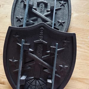 May include: Two black 3D printed shields with a sword and lightning bolts design. The shields are mounted on a metal stand.