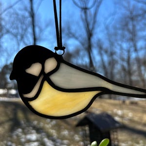 以下が含まれることがあります： 黒、白、黄色のガラス片でできたステンドグラスの鳥のオーナメント。鳥は黒い紐で吊り下げられています。