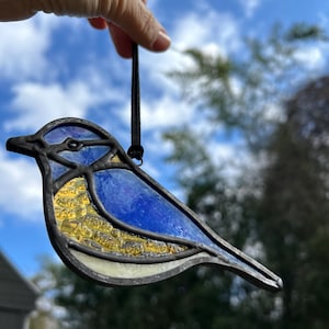 May include: A stained glass bluebird ornament with yellow accents hanging from a black string. The bird is suspended against a blue sky with white clouds.