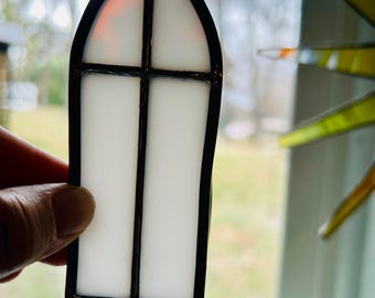 Könnte beinhalten: Ein Buntglas-Sonnenfänger in Form eines gotischen Spitzbogenfensters. Das Stück besteht aus undurchsichtigen weißen Glasscheiben, die durch einen schwarzen Metallrahmen in Kreuzform unterteilt sind, mit einer kleinen Metallöse zum Aufhängen.