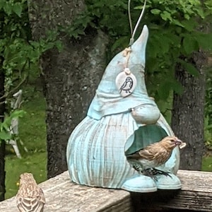 May include: A blue ceramic bird feeder shaped like a gnome with a small bird perched inside. The feeder has a white ceramic tag with a black silhouette of a bird on it.