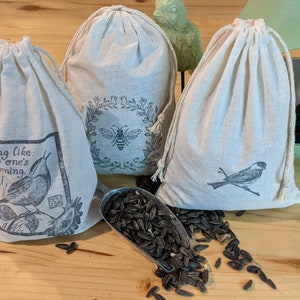 May include: Three white cotton drawstring bags with different bird and nature designs. The bags are filled with black birdseed. The text on one bag reads "Sing like no one's listening."