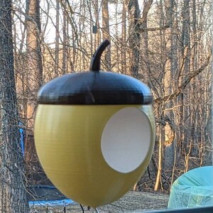 May include: A yellow acorn-shaped bird feeder with a brown top and a circular opening. The bird feeder is hanging in front of a window with a view of trees. The feeder is designed to attract birds to a garden or outdoor space.