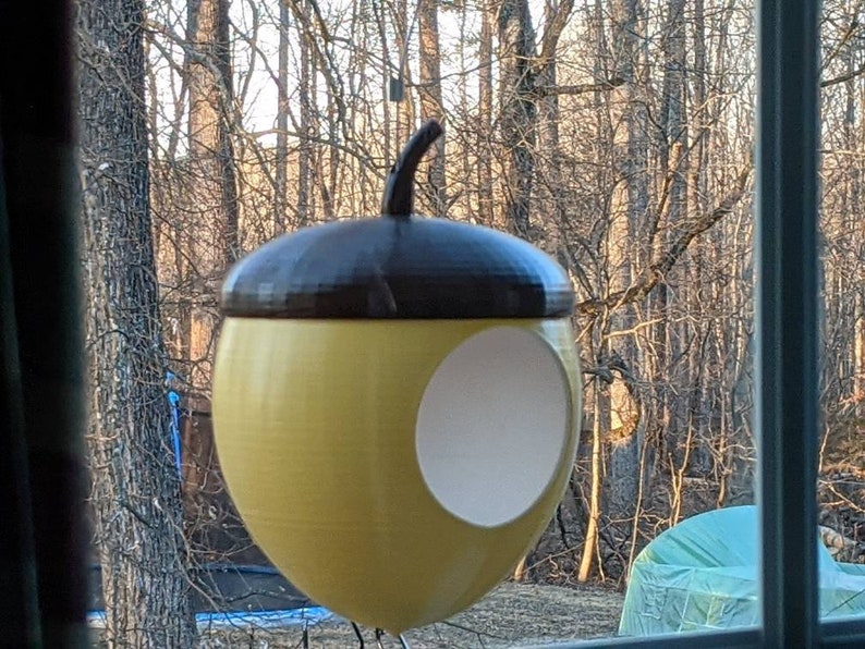 May include: A yellow acorn-shaped bird feeder with a brown top and a circular opening. The bird feeder is hanging in front of a window with a view of trees. The feeder is designed to attract birds to a garden or outdoor space.