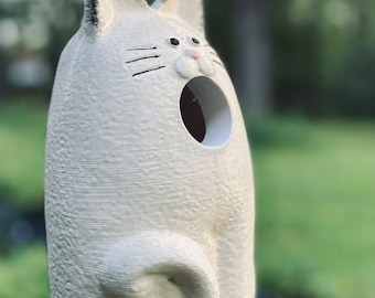 May include: White ceramic birdhouse shaped like a cat with a black eye and whiskers. The birdhouse has a round opening for birds to enter.