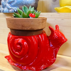 May include: A red snail planter with a brown terracotta pot inside. The pot is filled with a small succulent plant and a red and white mushroom figurine.