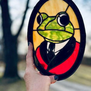 Op de afbeelding: Een gebrandschilderd raam met een afbeelding van een kikker in een zwart pak en een rode stropdas. De kikker heeft grote, ronde ogen en een groen lichaam. Het raam is ovaalvormig en heeft een zwart frame.