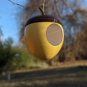 May include: A yellow and brown acorn-shaped birdhouse hanging from a tree branch. The birdhouse has a circular opening and a textured surface. The top is brown, and the body is yellow. The background is blurred, showing a blue sky and greenery.